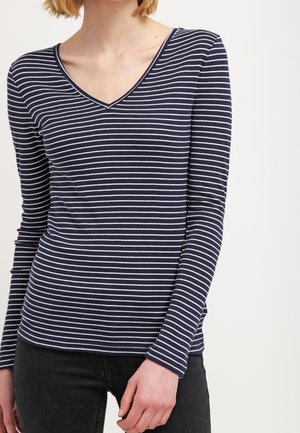 Frau trägt ein marineblaues Langarmshirt mit V-Ausschnitt mit dünnen weißen horizontalen Streifen und schwarzen Hosen vor einem schlichten Hintergrund.