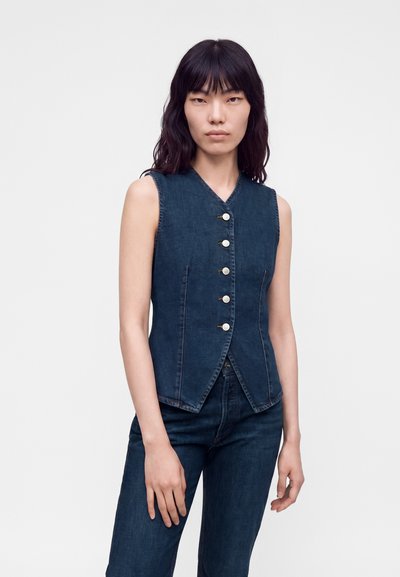 Femme aux longs cheveux foncés portant un gilet en denim bleu foncé sans manches avec des boutons blancs et un jean en denim bleu foncé assorti.