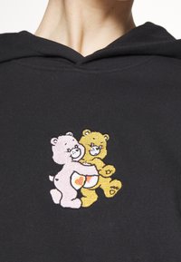 Sudadera negra con capucha que presenta un gráfico bordado de dos osos de dibujos animados, uno rosa y otro amarillo, sosteniendo un corazón, con una suave textura de algodón.