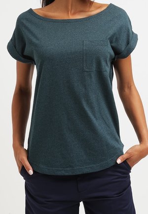Vrouw draagt een donkergroen kortärmelig topje met een klein borstzakje en marineblauwe broek, handen rusten in de zakken.