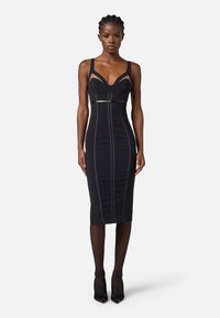 Abito bodycon nero con silhouette aderente, spalline larghe, scollatura a cuore e cuciture verticali a contrasto. Abbinato a scarpe con tacco alto.