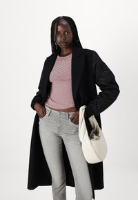 Een lange zwarte jas over een roodgestreepte top, grijze jeans en een crèmekleurige tas vasthoudend. De outfit heeft verschillende texturen en contrasterende kleuren.