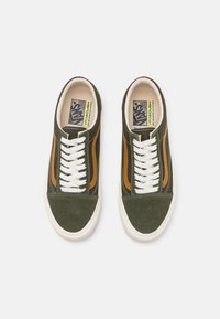 Zapatillas de ante verde con cordones blancos, que presentan una franja de acento marrón y una suela de goma blanca. Etiqueta visible dentro del zapato.