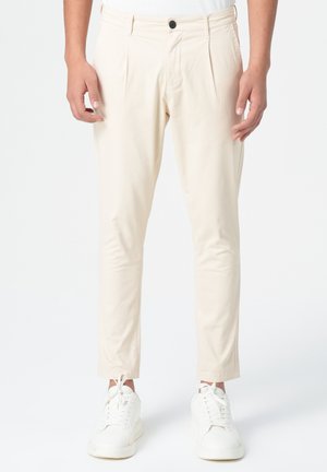 Pantalons sur mesure couleur crème avec deux plis à l'avant, fermeture éclair et un seul bouton. Associés avec des baskets blanches.