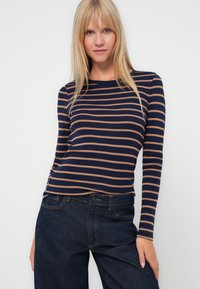 Femme aux cheveux blonds raides portant un t-shirt à rayures bleu marine et beige à manches longues et un jean bleu foncé, se tenant devant un fond clair et uni.