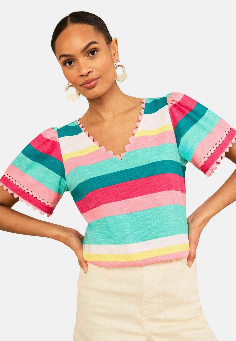 Frau mit dunklen Haaren und Ohrringen, die ein bunt gestreiftes Kurzarmshirt mit gewelltem Ausschnitt und helle Hose trägt, Hände in der Hüfte.