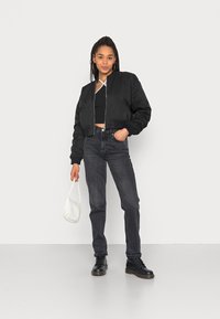 Jaqueta bomber cropped preta com punhos canelados, combinada com jeans escuros de cintura alta e botas pretas com atacadores. Bolsa branca complementa o visual.