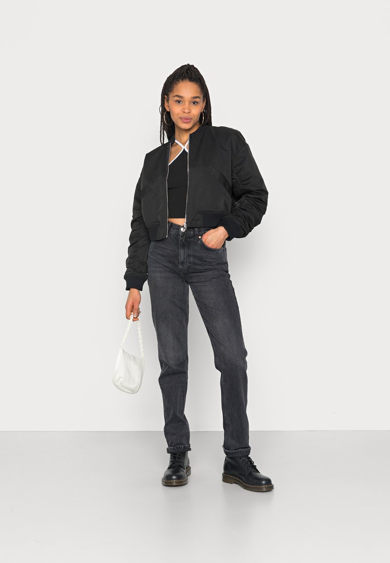 Jaqueta bomber cropped preta com punhos canelados, combinada com jeans escuros de cintura alta e botas pretas com atacadores. Bolsa branca complementa o visual.