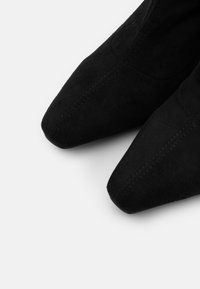 Botas de ante negras de tacón y punta afilada, con una textura suave y costuras decorativas a lo largo de las costuras. Diseño simple y elegante.