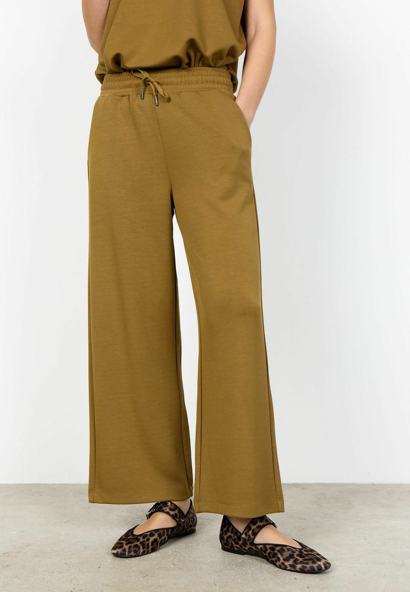 Pantalones anchos verde oliva con cintura elástica y cordón, combinados con zapatos Mary Jane de estampado de leopardo sobre un suelo gris.