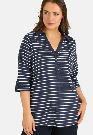 Vrouw draagt een donkerblauw-wit horizontaal gestreept overhemd met kraag, driekwart mouwen en knoopdetails, staand met één hand in de zak.