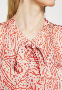 Hedvábná blůza s jemně krémovým podkladem a červenými abstraktními vzory, v výstřihem do V a s nařaseným detailem zavazování na límci.