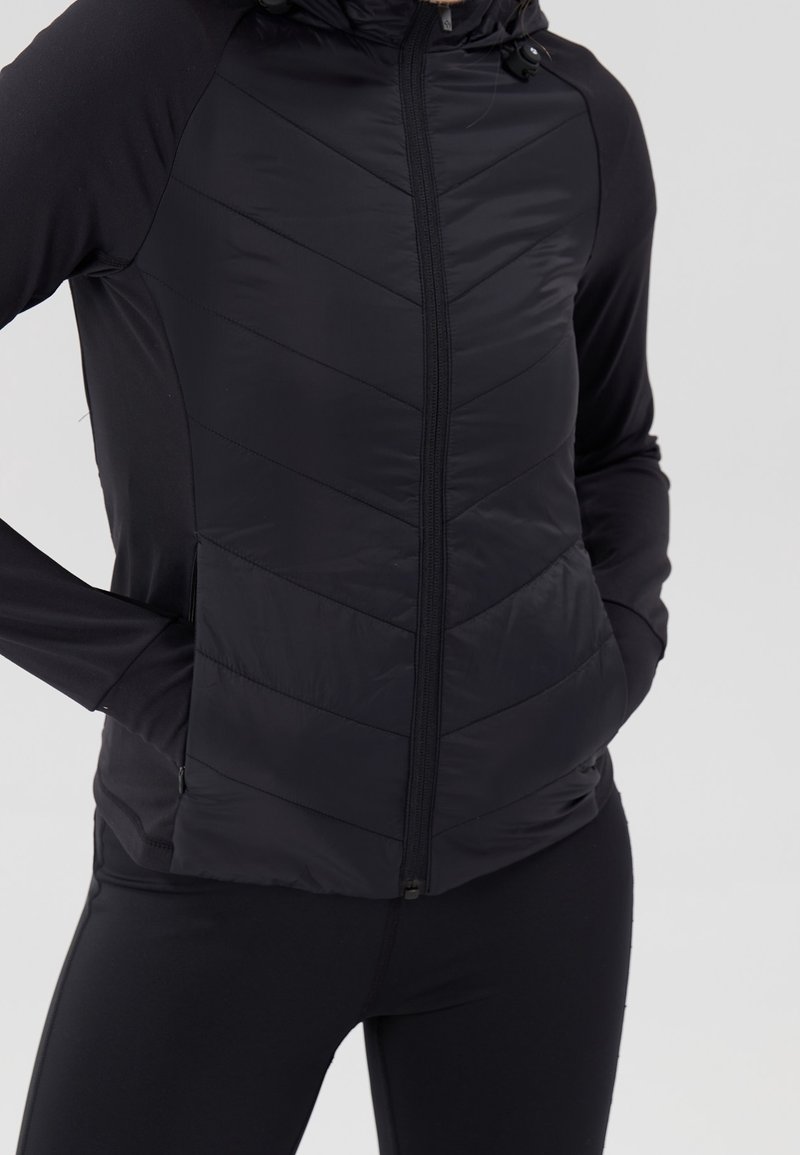 Chaqueta negra con cremallera hecha de material suave e aislante con costuras horizontales en forma de chevron. Cuenta con capucha y bolsillos laterales, mangas largas.