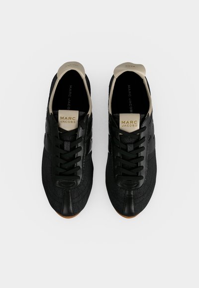 Paire de baskets noires Marc Jacobs avec des languettes de talon beige et des étiquettes logo sur la languette, vue de dessus sur fond blanc.