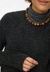 Mörkgrå texturerad tröja med långa ärmar, utrustad med ribbad mudd, bärs över en grå polotröja och accentueras av ett pärlhalsband.