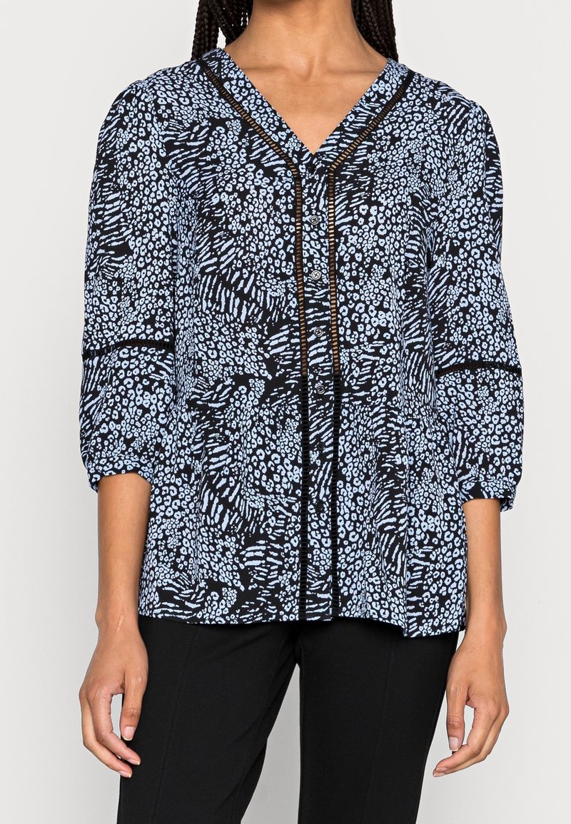 Femme portant un chemisier noir et bleu clair à motif avec col en V, manches trois-quarts et pantalon noir.