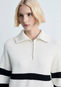 Weißer Strickpullover mit einem Reißverschlusskragen, der strukturierte Streifen und schwarze horizontale Akzente aufweist. Entspannter Schnitt und gerippte Details.