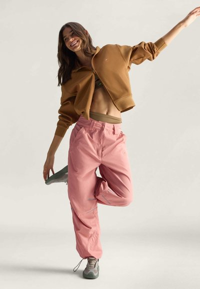 Brauner Reißverschluss-Sweatshirt, pinke lockere Hose und graue Sneakers. Sanfte Materialien mit leichtem Glanz; die Hose verfügt über seitliche Taschen und reflektierende Akzente.