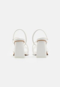 Sandales à talons blocs blanches avec une texture lisse, dotées de brides croisées et d'une sangle autour de la cheville pour un maintien sécurisé. Design simple et moderne.