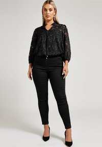 Blouse noire transparente avec broderie florale, associée à un jean skinny noir et des chaussures à talons hauts noires. Des accessoires minimalistes complètent le look.