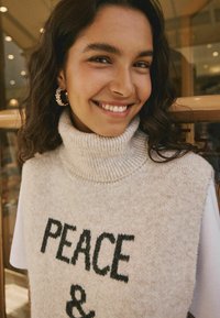 Maglione beige a collo alto con "PEACE &" ricamato in lettere nere. Presenta un motivo a maglia morbido e testurizzato, indossato su una camicia bianca.