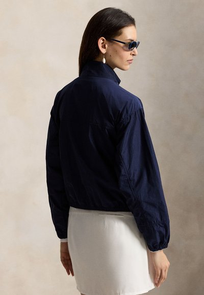 Marineblå lett jakke med opprett krage og elastiske mansjetter, kombinert med et hvitt skjørt. Stoffet virker glatt og lett teksturert.