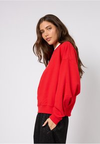 Červená široká mikina s kulatým výstřihem a žebrovanými manžetami, doplněná volnými černými kalhotami s jemným vzorem.