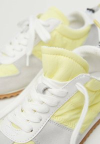 Gros plan de baskets blanches, jaunes et grises avec des lacets blancs et des languettes rembourrées douces sur un fond clair.