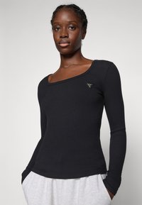 Woman wearing black long-sleeve ribbed top with triangular logo on chest, paired with light gray pants, standing against a plain background.