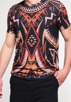 Homme portant un T-shirt noir à manches courtes avec des motifs géométriques symétriques dans des tons marron, rouge et beige.
