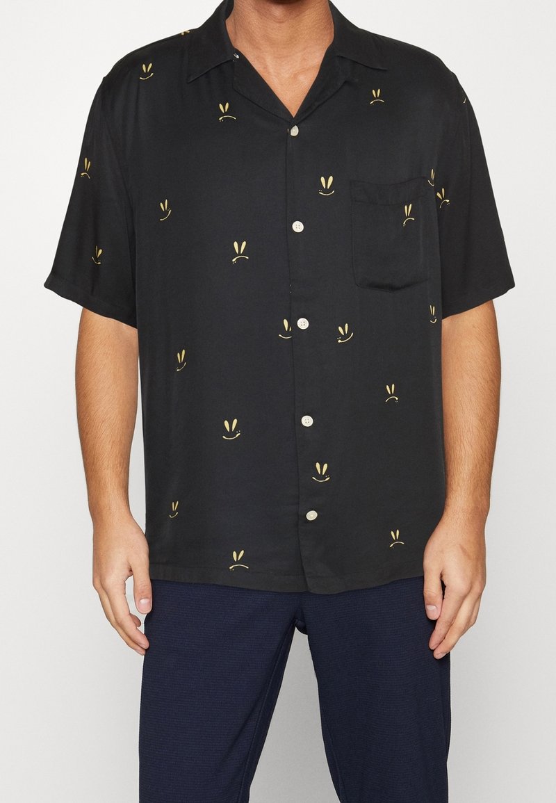 Chemise noire à manches courtes avec boutons, motifs de petits visages souriants jaunes et une poche poitrine gauche, portée avec un pantalon bleu foncé.