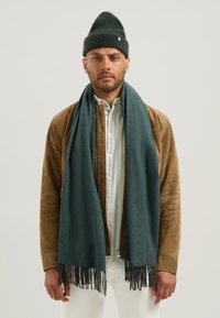 Muž v tmavozelené pletené čepici, odpov�ídajícím dlouhém třásňovém šálu, hnědé semišové bundě a bílé košili s knoflíky stojí proti jednobarevnému pozadí.