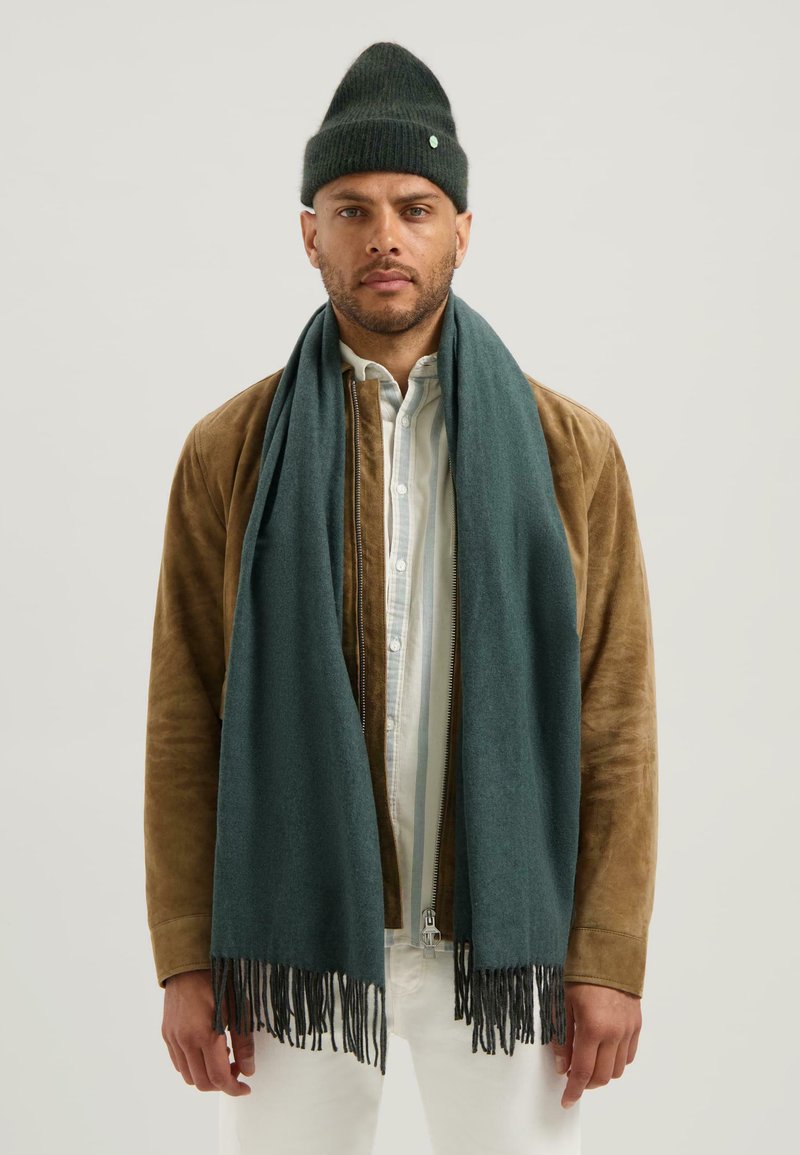 Muž v tmavozelené pletené čepici, odpovídajícím dlouhém třásňovém šálu, hnědé semišové bundě a bílé košili s knoflíky stojí proti jednobarevnému pozadí.