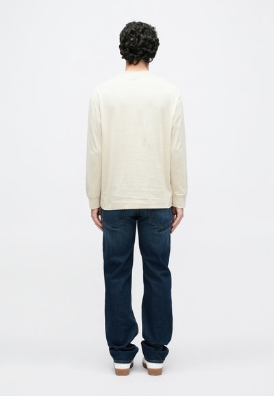 Gaiši bēša garām piedurknēm krekls ar apaļu apkakli, kombinēts ar tumši zilām džinsa biksēm un kurpēm ar brūnu zoli. Gluda auduma tekstūra, ikdienišķa piegriezuma.