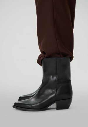 Zwarte leren enkelboots met lage blokhakken, gedragen met donkerbruine broeken.
