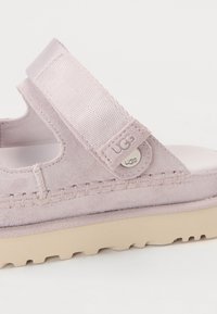 Sandale en daim violet clair avec sangle ajustable, accent métallique UGG de marque, semelle épaisse beige et détails surpiqués sur le côté.