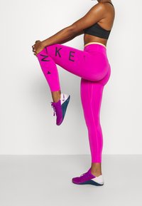 Leggings rosa con testo "NIKE" nero e accenti geometrici. Realizzati in materiale elasticizzato. Abbinati a scarpe da ginnastica multicolori.