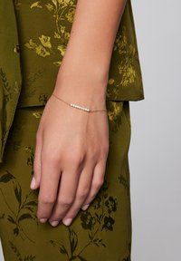 Złota bransoletka z cienkim łańcuszkiem, ozdobiona rzędem małych, przezroczystych kamieni szlachetnych. Noszona na dłoni na tle oliwkowo-zielonego materiału w kwiatowy wzór.