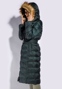 Green puffer coat with a fur-lined hood, belted waist, long length, quilted texture, and side zip pockets.