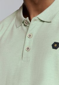 Lichtgroen poloshirt van een gestructureerde stof, met een twee-knopen sluiting en een zwart hexagonaal logo label.