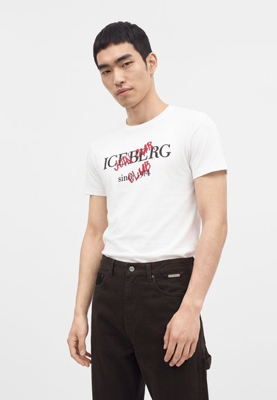 Νέος άνδρας που φοράει λευκό T-shirt με μαύρο και κόκκινο κείμενο σε συνδυασμό με μαύρο παντελόνι μπροστά από απλό ανοιχτό φόντο.