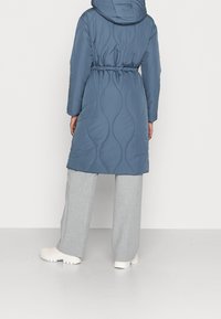 Abrigo azul acolchado con capucha, que presenta un patrón de costura ondulado y cintura con cordón, combinado con pantalones de pierna ancha en gris claro y zapatos blancos.