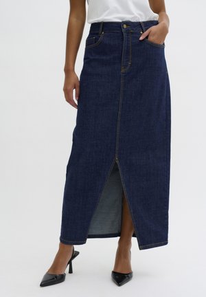Jupe maxi en denim bleu foncé avec une fente avant, des coutures claires et des poches latérales ; associée à des escarpins noirs à bout pointu.
