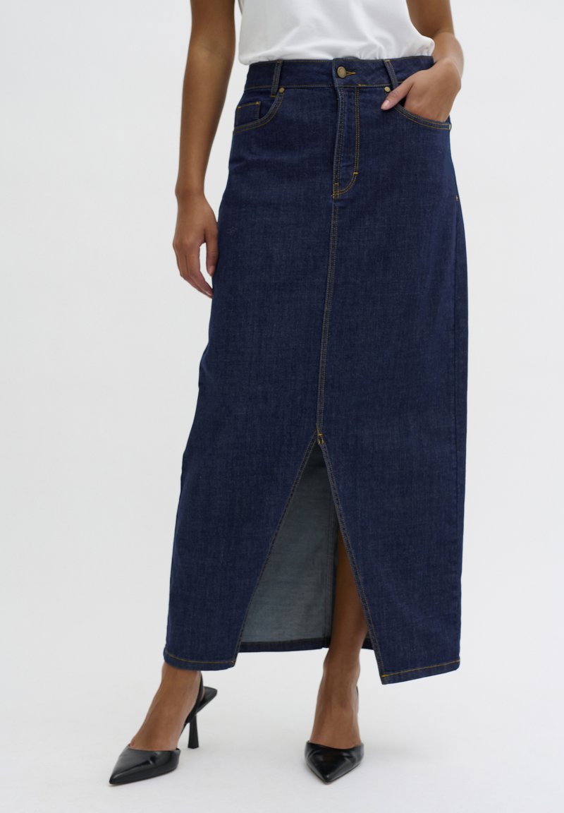 Gonna maxi in jeans blu scuro con spacco frontale, cuciture chiare e tasche laterali; abbinata a tacchi a punta neri.
