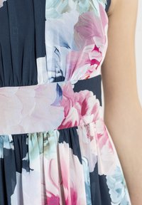 Robe florale avec une base en tissu bleu marine ornée de motifs de fleurs roses claires, bleues et blanches. La ceinture crée des plissés froncés au niveau de la jupe.