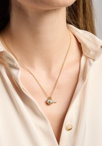 Collar de cadena de oro con un pequeño colgante de ave colorida con acentos azules y verdes, descansando sobre una blusa beige.