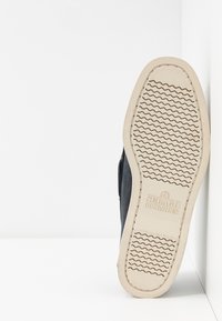 Black slip-on shoe with rubber sole, featuring a textured wave pattern and embossed logo on the underside. Neutral color insole contrasting design.