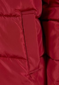 Giacca a vento rossa con finitura liscia e lucida, design compatto e tasca laterale. Il tessuto appare morbido e isolato.