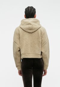 Κοντή μπεζ φ�όρμα από αφράτο, υφασμάτινο υλικό; διαθέτει μεγάλη κουκούλα και φαρδιά μανίκια. Συνδυάζεται με σκούρα καφέ παντελόνια.