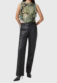 Femme portant un haut sans manches vert à motifs floraux avec des côtés froncés, un pantalon en cuir noir taille haute et des bottes à talons aiguilles noires.